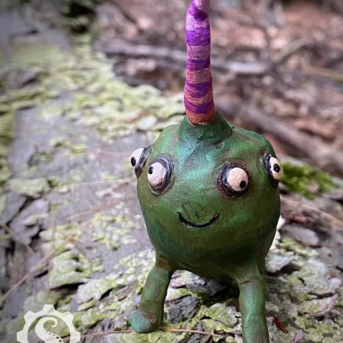 Silly, smiling green creature with four bulging eyes looking in all different directions. A long pink and purple striped horn extends from the top of its head.