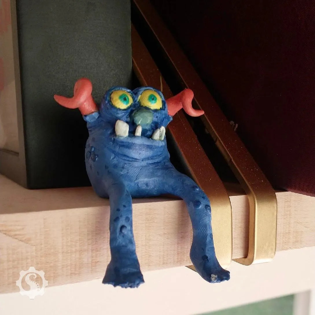 An original character sculpture of a small monster figure that sits ona shelf with his legs hanging over..